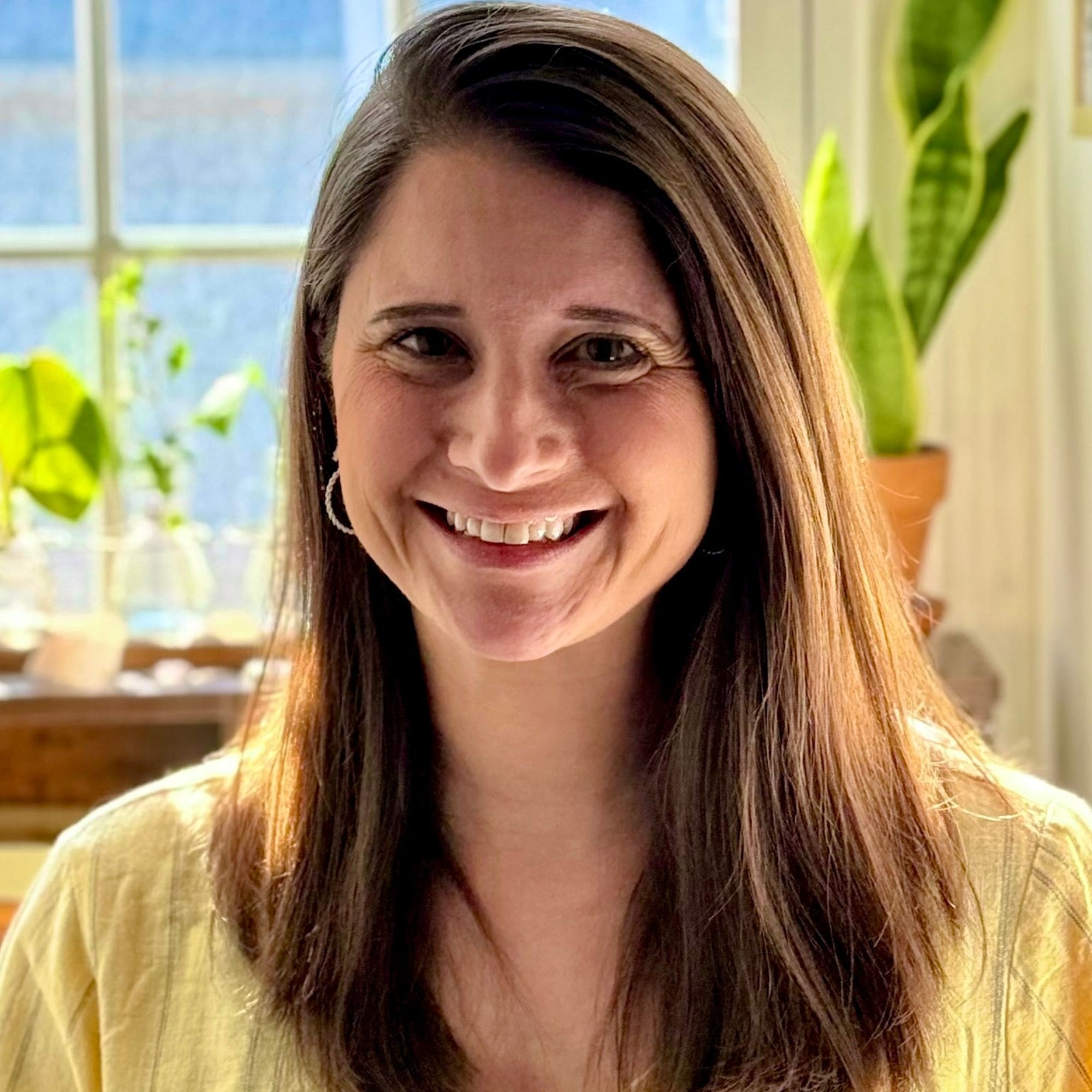 **Alt text:** Close-up headshot of a person with long brown hair smiling at the camera, softly lit by daylight, with green houseplants and a sunlit window in the background.