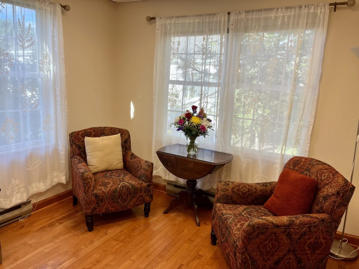 Two patterned armchairs around a small drop-leaf table with a vase of flowers by large windows.