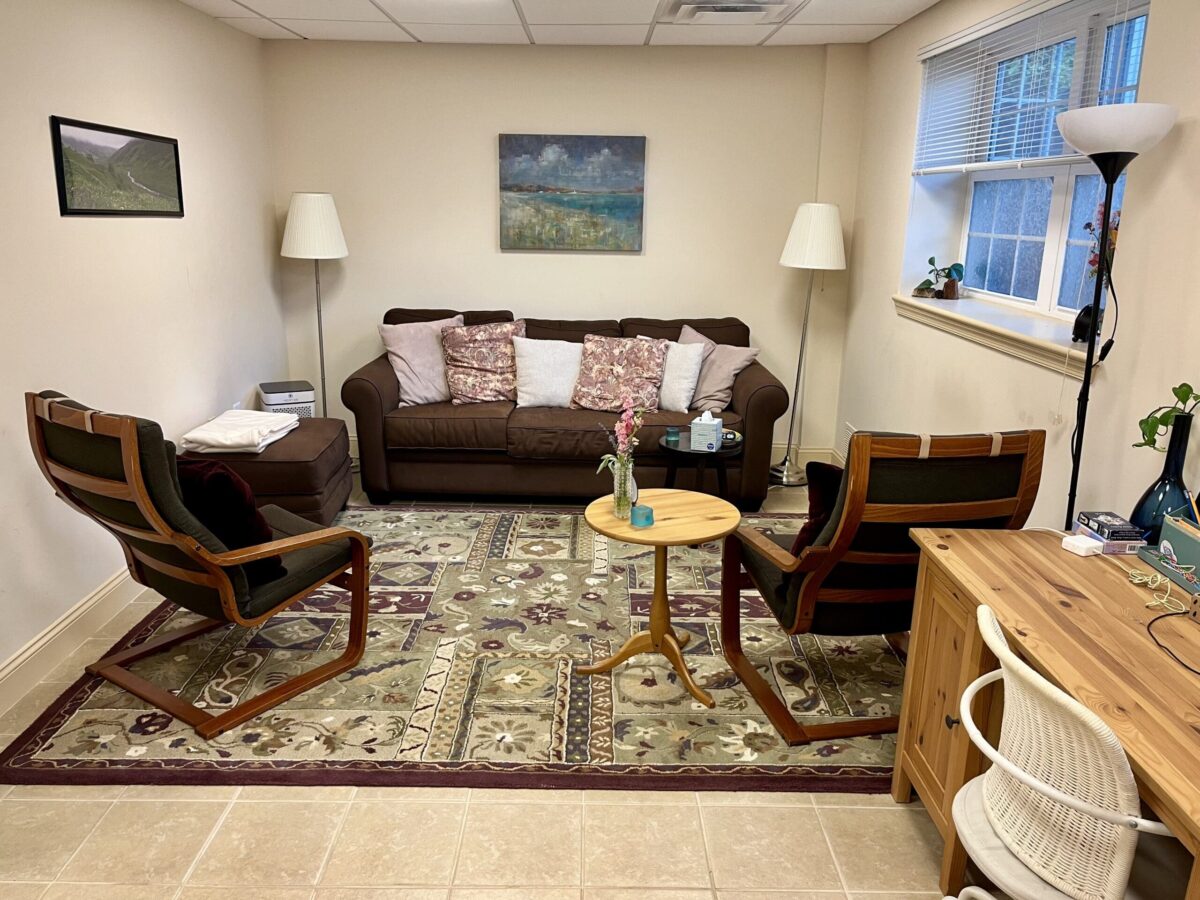 Office with brown sofa, matching chairs, throw pillows, two floor lamps, coffee table, area rug, wall art, window with blinds, and wooden desk with chair.